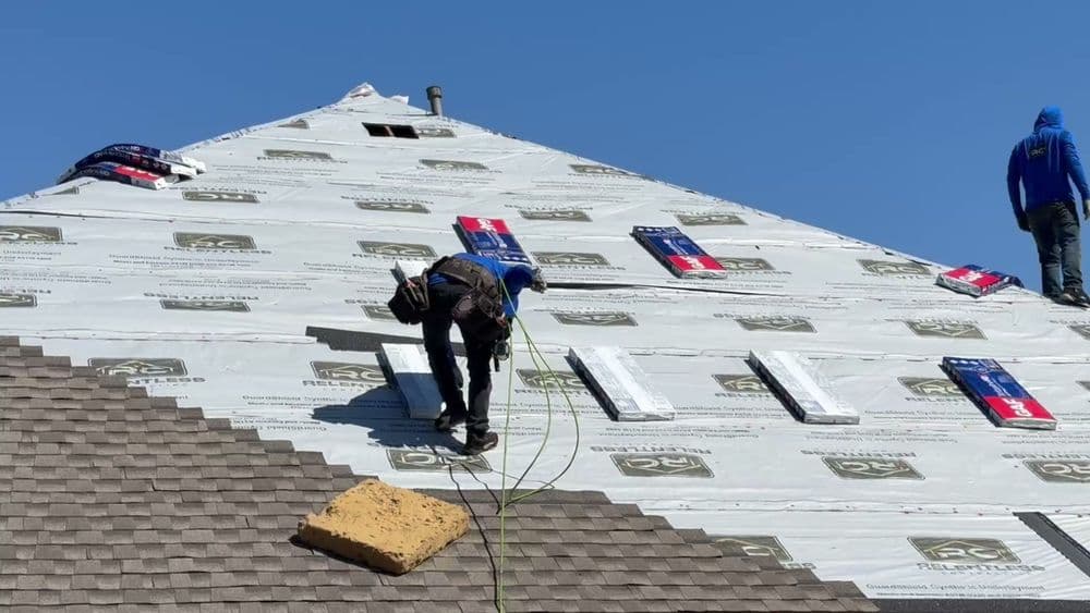 Roofing workers installing shingles on a house under a clear blue sky. Safety harnesses visible.