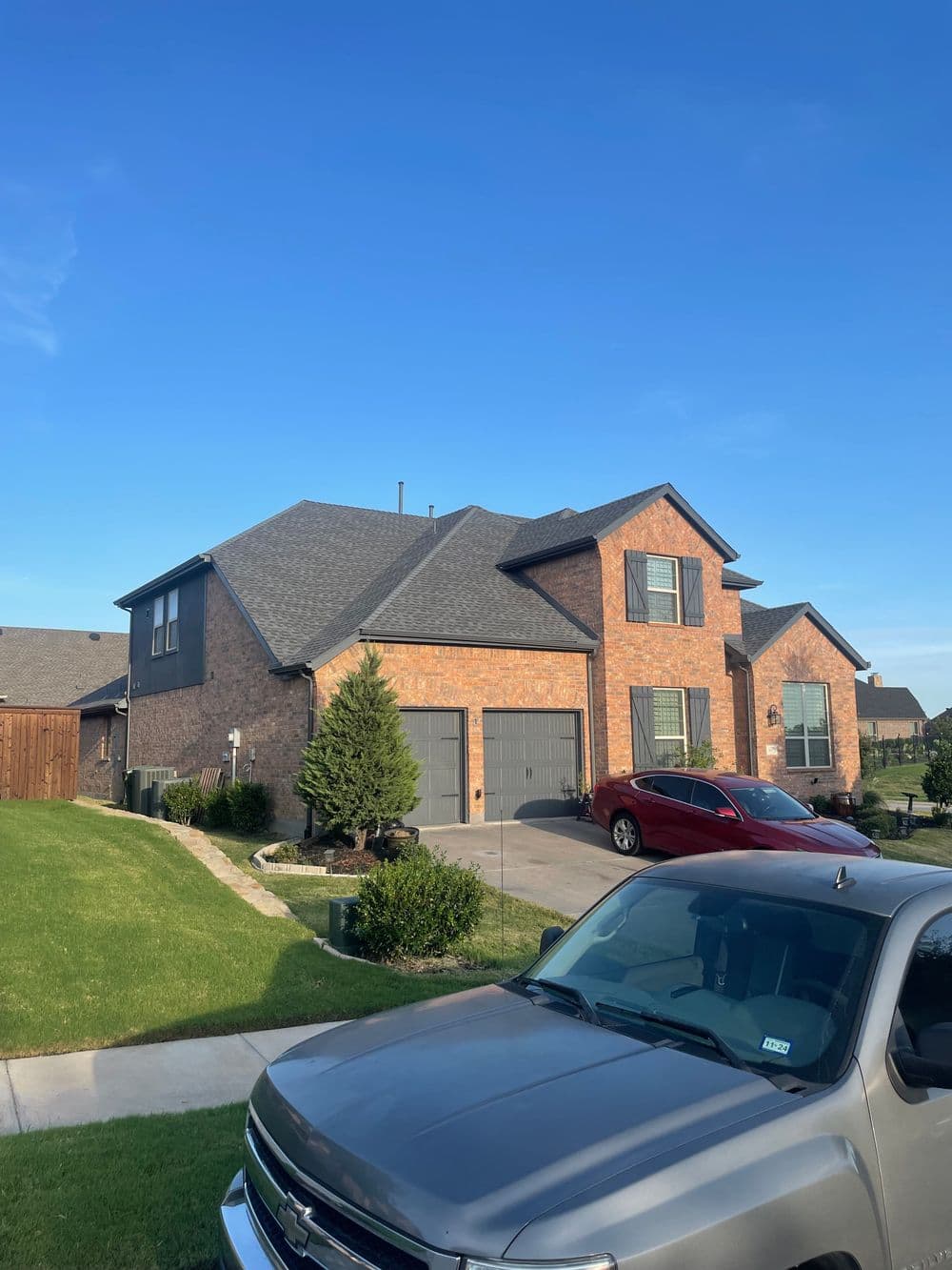 Brick suburban house with a gray roof, green lawn, and parked cars in the driveway.
