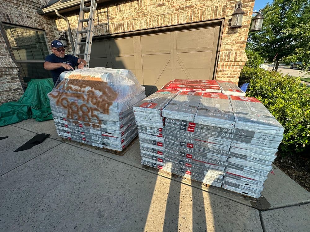 A man unpacking pallets of roofing shingles in front of a house.