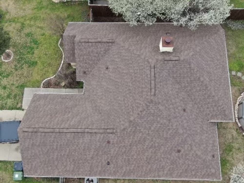 Aerial view of a house roof with asphalt shingles and chimney, surrounded by a green yard.