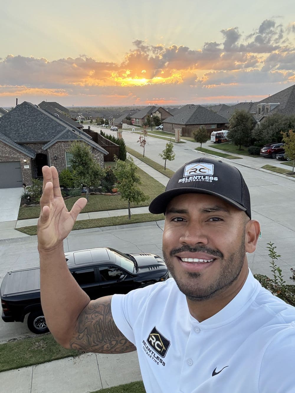 Man in a branded polo shirt waving at sunset over suburban neighborhood.