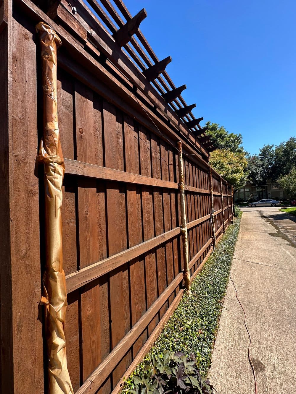 Wooden fence installation along a sunny pathway, with greenery below and clear blue sky.