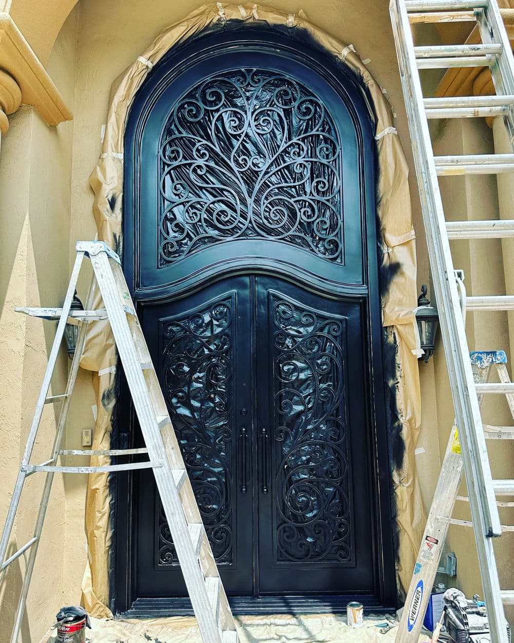 Black ornate double door with decorative ironwork, surrounded by ladders and painter's tape.