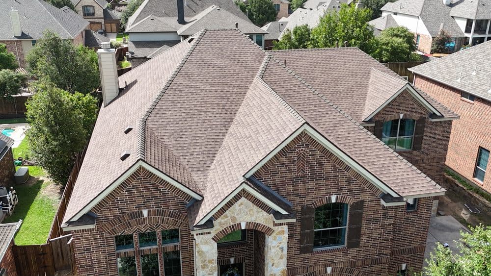 Aerial view of a suburban brick house with a tan shingle roof and landscaped yard.