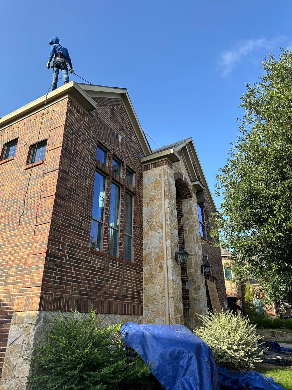 Roof maintenance on a brick house with a worker harnessed on top and a blue tarp below.