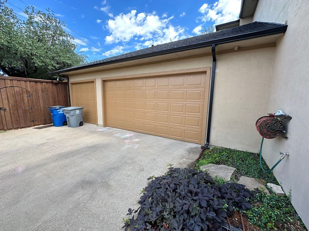 Garage with beige doors, blue and gray trash bins, and landscaping in a sunny outdoor setting.