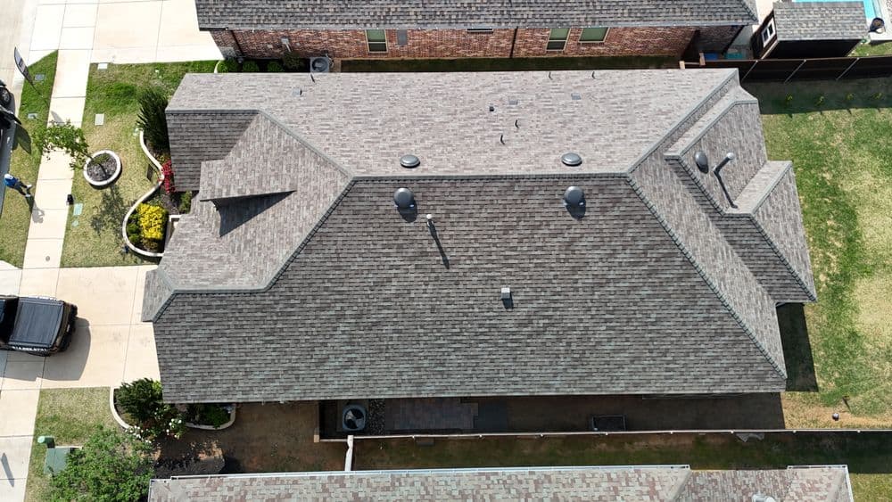 Aerial view of a residential roof with shingles and ventilation, showcasing landscaping.
